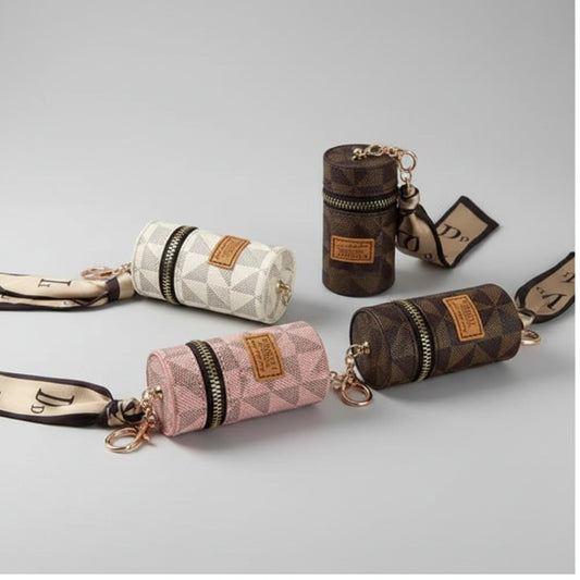 Three cylindrical zippered pouches with geometric monogram patterns in pink, cream, and brown, each with a matching neck scarf keychain and gold-tone keyring, styled on top of printed papers alongside a white earbud case and lipstick.