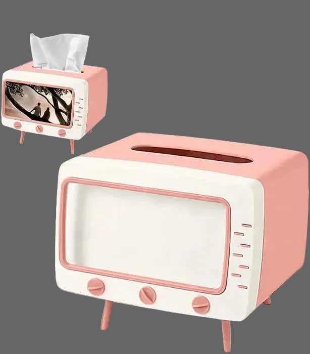 A retro-style tissue box cover or small dispenser shaped like a television, shown in pink and white on a wooden surface. A second matching box sits behind it on a wood block against a dark gray wall.